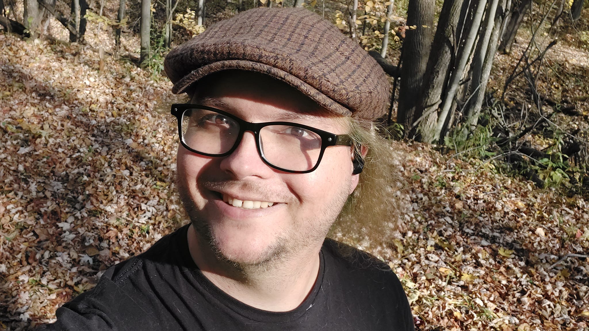 A man is wearing a brown flat cap and black shirt, standing on a forest path